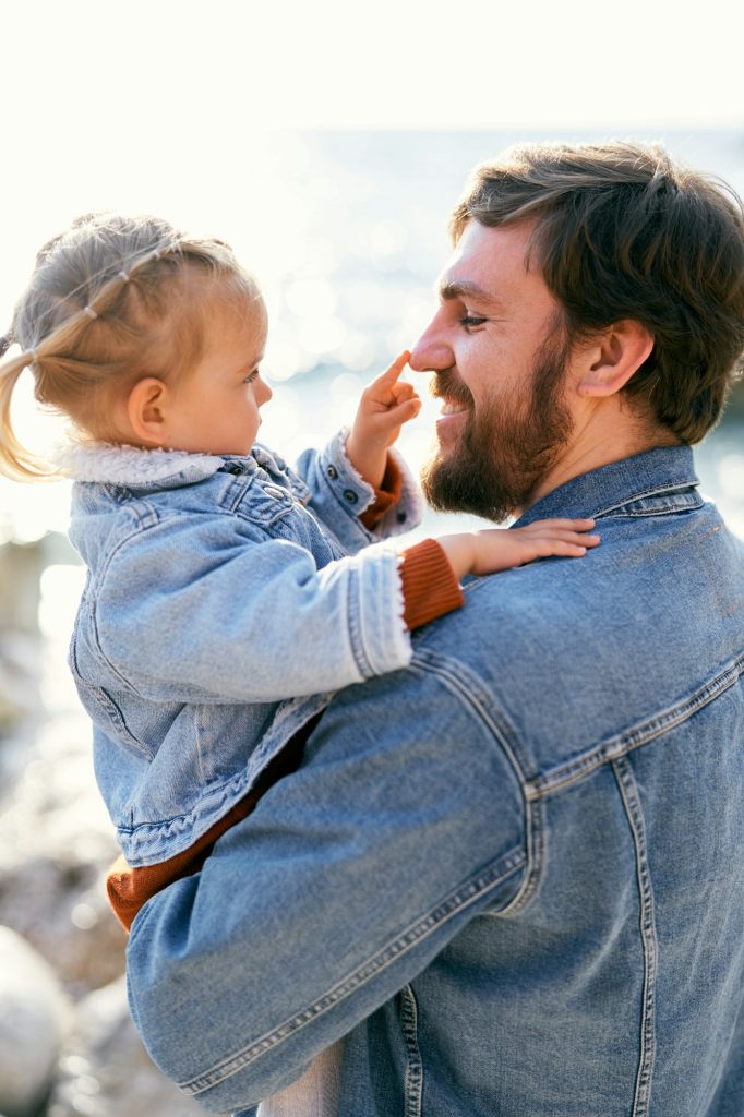 Smiling dad is holding a little girl in his arms, who touches his nose with a finger. Close-up